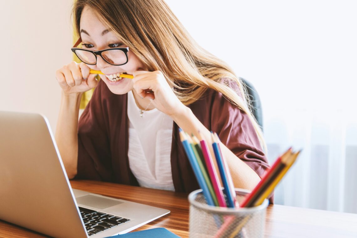 A person wearing glasses bites a pencil while looking at a laptop; colored pencils and a notebook are placed on the desk.