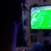 A person watches a soccer game on a wall-mounted TV in a dimly lit room.