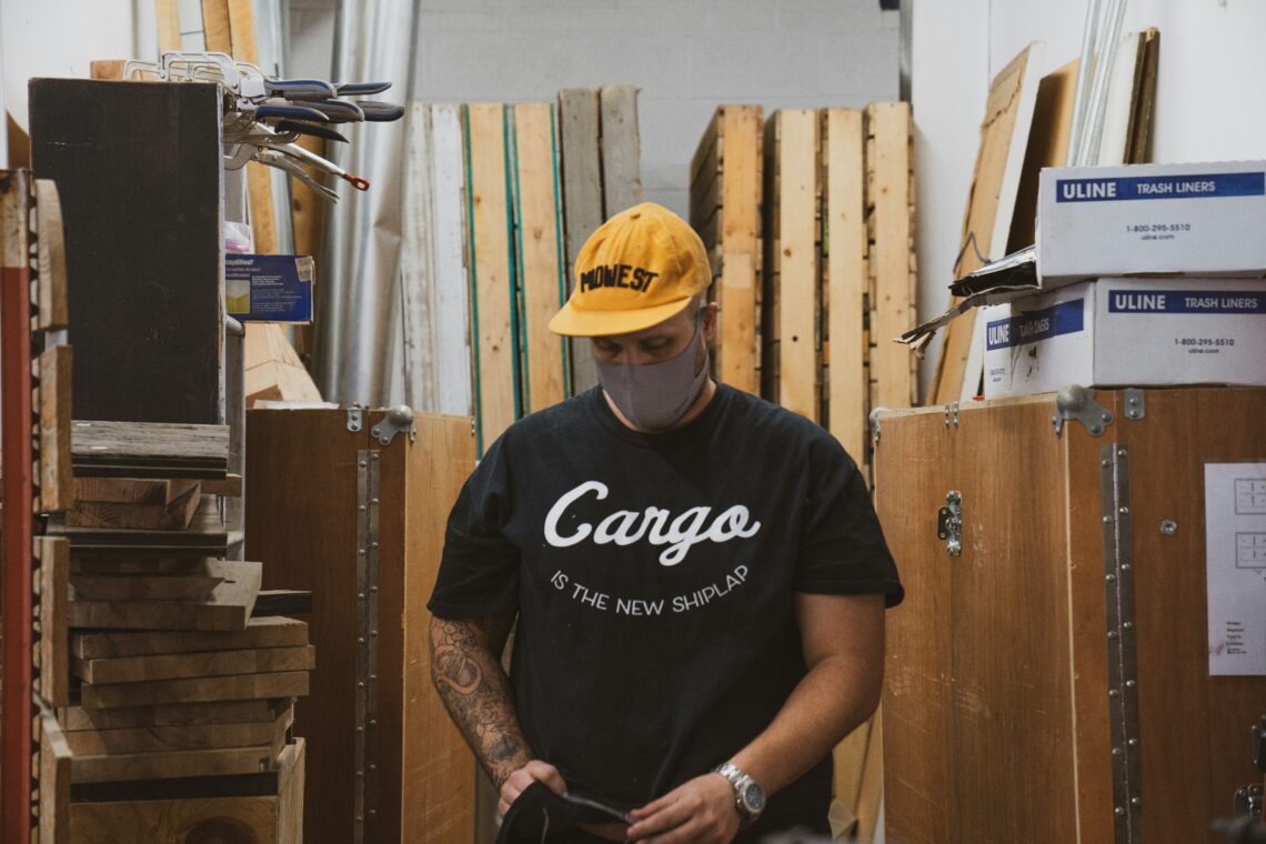 Person in a black "Cargo" shirt and yellow hat standing in a storage room.