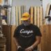 Person in a black "Cargo" shirt and yellow hat standing in a storage room.